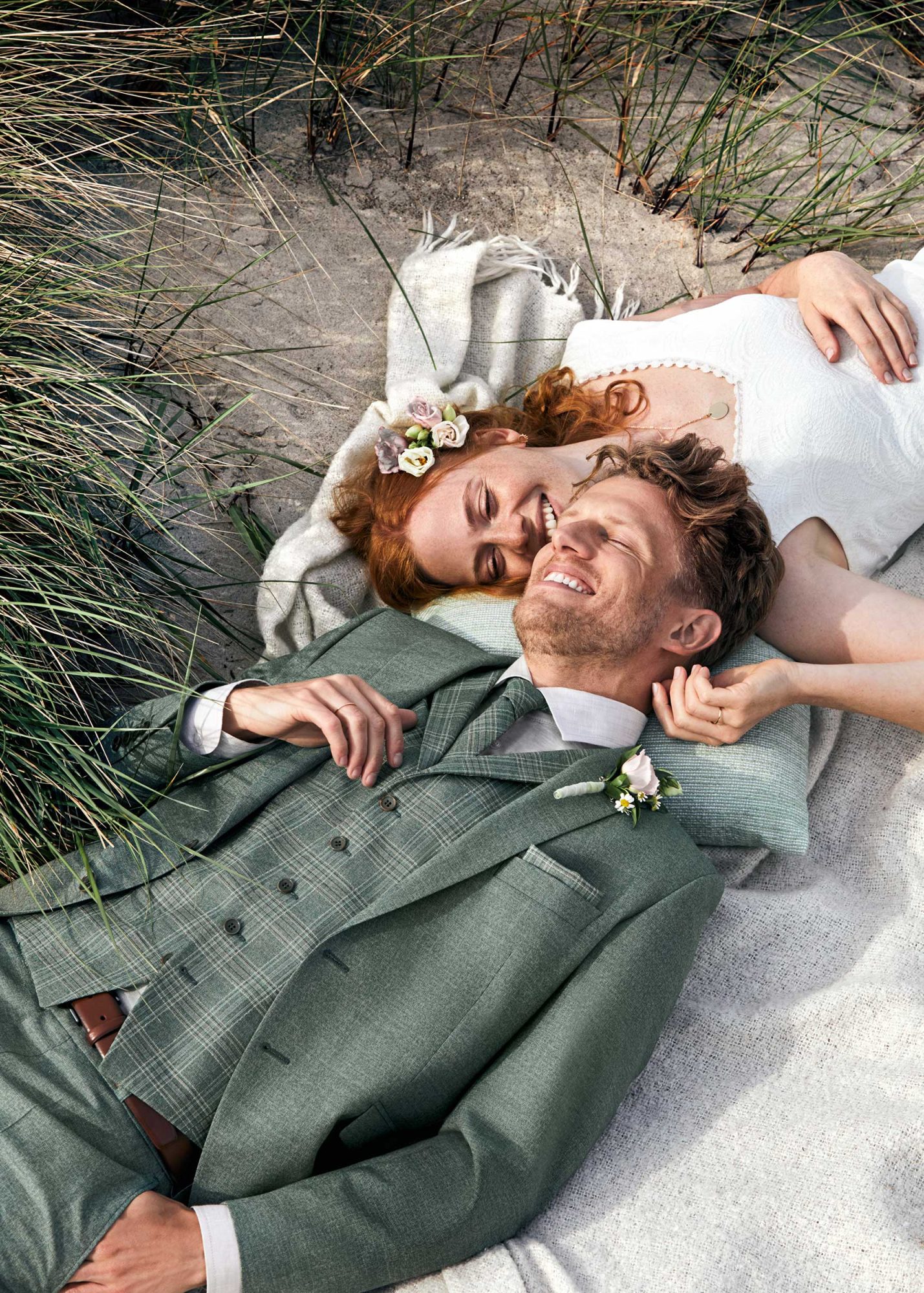 Brautpaar liegt am Strand in Hochzeitskleidung. Mann trägt grünen, karierten Anzug.