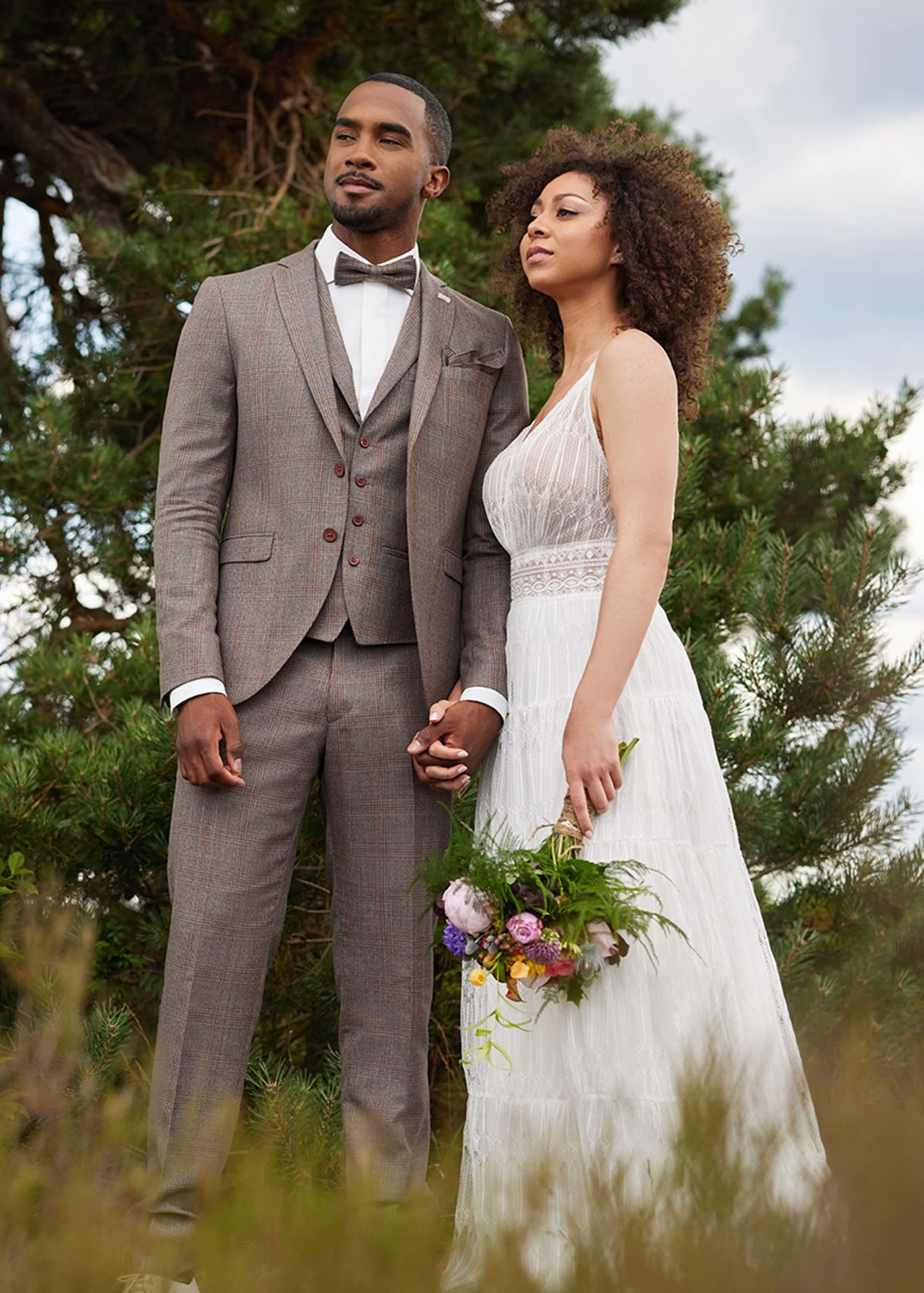 Brautpaar in der Natur. Bräutigam trägt einen braunen, karierten Anzug. Braut trägt ein schlichtes Hochzeitskleid.